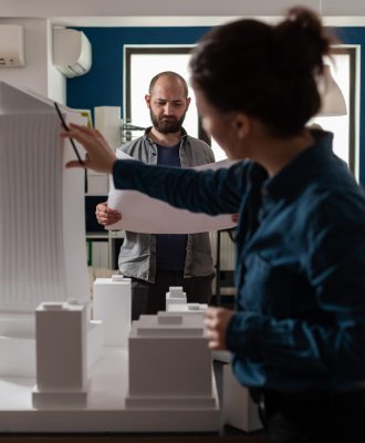 Architecture workers checking blueprints with maquette building model design plan. Caucasian workmates pointing at construction project layout for urban development business