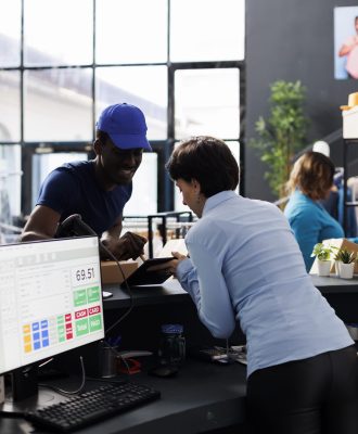 African american courier signing distribution document on tablet computer, discussing customers shipping details with store manager in modern boutique. Employee preparing packages in shopping centre