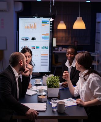 Workaholics businesspeople brainstorming financial company ideas analyzing strategy paperwork late at night in business office meeting room. Multi-ethnic coworkers checking solution